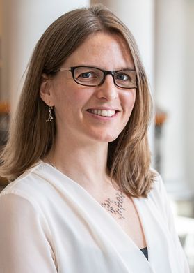 Headshot of WVU Extension professor Jennifer Robertson Honecker. She is standing inside with white columns in the background. She is wearing a white blouse and has shoulder length blonde hair. She is wearing black-rimmed glasses. 