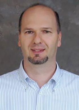 WVU professor Gianfranco Doretto is pictured here in front of a gray marble background wearing a blue pin striped shirt. He has dark hair and a dark goatee. 