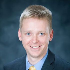 Headshot of WVU Lane Innovation Hub director Dustin Spayde. He is pictured against a blue background wearing a navy blue sport coat over a light blue dress shirt and a yellow tie. He has short blonde hair. 