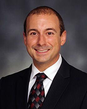 Headshot of WVU Pharmacy assistant professor Mark Garofoli. He is pictured against a gray background and is wearing a dark colored suit with a white dress shirt and red and black checkered tie. He has thinning dark hair. 