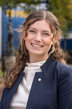Headshot of WVU student Sophia Flower. She is pictured outside wearing a navy blue jacket over a white turtleneck shirt. She has curly, long blonde hair. 