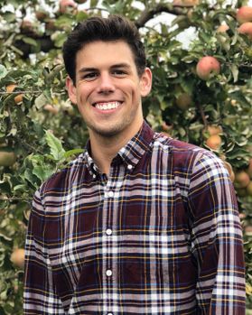 Headshot of WVU clinical psychology graduate student Johnathan Meier. He is pictured standing in front of a green apple tree with ripe red apples behind him. He is wearing a maroon and white flannel shirt and has short brown hair. 