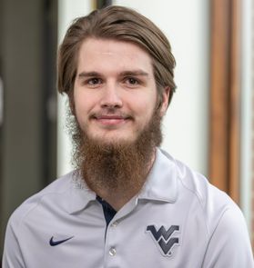 Headshot of WVU student and Mountaineer mascot candidate Braden Adkins. He is pictured wearing a gray WVU golf shirt. He has light brown hair and a long, light brown beard. 