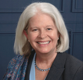 Headshot of WVU researcher Marian Reven. She is pictured against a blue gray background and is wearing a dark gray jacket over a light blue shirt with a patterned scarf. She has shoulder length gray hair and is wearing a blue necklace and earrings. 