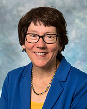Headshot of WVU researcher Kathryn Moffett. She is pictured against a mottled blue background and is wearing a royal blue jacket over a gold blouse. She has short brown hair and wears dark-framed glasses. 
