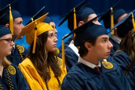 A graduate sits and listens in a gold cap and gown. Other graduates are shown surrounding her in blue caps and gowns.
