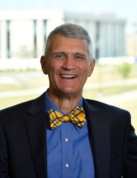 A person wears a dark jacket, blue shirt and gold and blue tie. Part of the WVU Evansdale Campus is visible in the background.