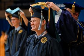 A graduate wearing a blue cap and gown smiles while being hooded during the College of Applied Human Sciences ceremony.