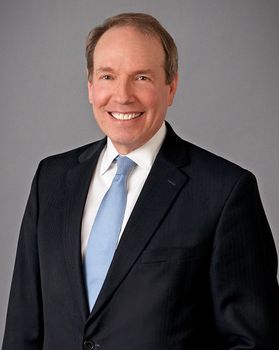 Headshot of lawyer Thomas Heywood. He is pictured in front of a gray background wearing a dark suit, white dress shirt and light blue tie. He has short, light brown hair. 