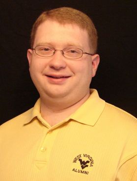Headshot of WVU award recipient Chad Proudfoot. He is pictured against a dark background wearing a yellow golf shirt with "West Virginia alumni" embroidered on the left chest area. He has red hair and wears glasses. 