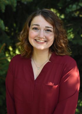Headshot of WVU student Anastasia Lucci. She is pictured outside with green trees behind her. She is wearing a burgundy blouse and has shoulder length auburn hair.