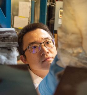 Headshot of Wenyuan Li. He is pictured inside of his lab closely examining an object. he has black hair and wears glasses. 