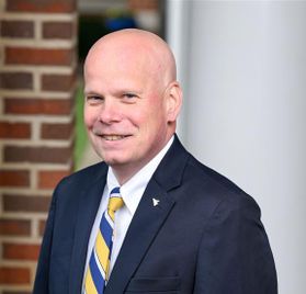 Headshot o WVU employee Kevin Berry. He is pictured outside with a red brick wall and white column behind him. He is wearing a navy blue coat over a white dress shirt with a blue and gold striped tie. He is bald. 