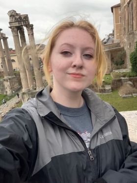 Headshot of WVU student Shelby Meador. She is pictured outside standing in front of ancient ruins. She is wearing a black and gray raincoat and has her blond hair pulled back. 