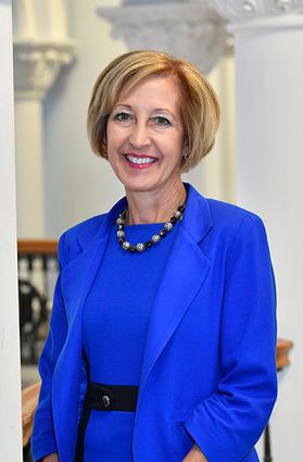 West Virginia Public Education Collaborative Director Donna Peduto is pictured standing inside with large white columns behind her. She is wearing a royal blue jacket and dress with a large bauble necklace. She has short blonde hair. 