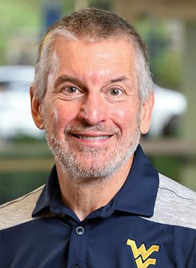 Headshot of WVU researcher Brad Humphreys. He is pictured wearing a WVU branded golf shirt that is navy blue and gray. He has short gray hair and a gray beard. 