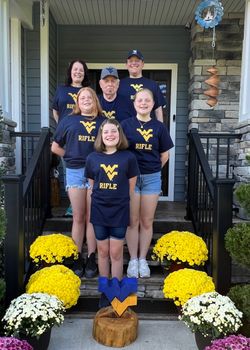 A photograph of the Hayhurst family all wearing the same WVU rifle T-shirt. The six family members are standing on their front porch surrounded by white, yellow and pink mums in full bloom and a wooden carving of the Flying WV logo.