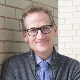 Headshot of WVU researcher Duncan Lorimer. He is standing against a white brick wall wearing a gray sweater over a blue checked shirt and lavender tie. He has medium, light colored hair and wears black framed glasses. 