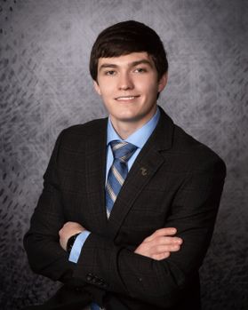 Photo of smiling young man with dark hair, dark suit, blue shirt and tie, arms crossed in front