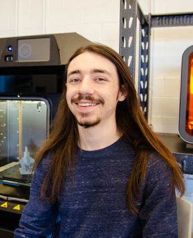 Headshot of WVU student Dylan Printy. He is pictured inside with industrial machines behind him. He is wearing a navy blue sweater and has long brown hair and a brown goat tee. 