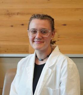 Headshot of WVU student Luna Martin. She is pictured inside wearing a white lab coat over a black shirt. She has braided blonde hair and wears glasses. 