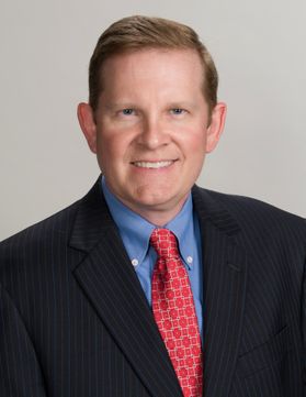Headshot of WVU honoree Loren Lazear. He is pictured in front of a beige background wearing a black coat over a light blue shirt with a red patterned tie. He has short, light brown hair. 
