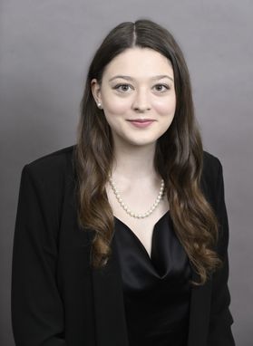 Headshot of WVU researcher Angelyn Gemmen. She is pictured against a greige background and is wearing a black jacket over a shiny black blouse. She has long, curled dark hair and fair skin. 