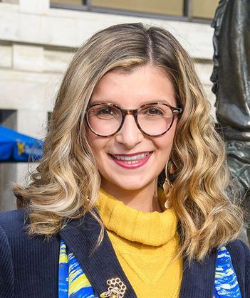 Headshot of WVU Fulbright Scholar Giana Loretta. She is pictured outside with a light colored building in the background. She is wearing a gold turtleneck sweater under a navy blur blazer with a blue and gold scarf. She has shoulder length blonde hair. 