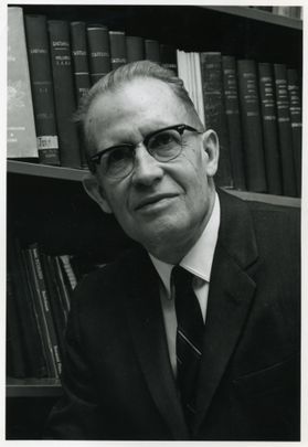 A headshot of WVU researcher Earl Core. The photograph is black and white and shows Core, dressed in a dark suit, white dress shirt and dark striped tie, standing in front of stacks of books on shelves. 