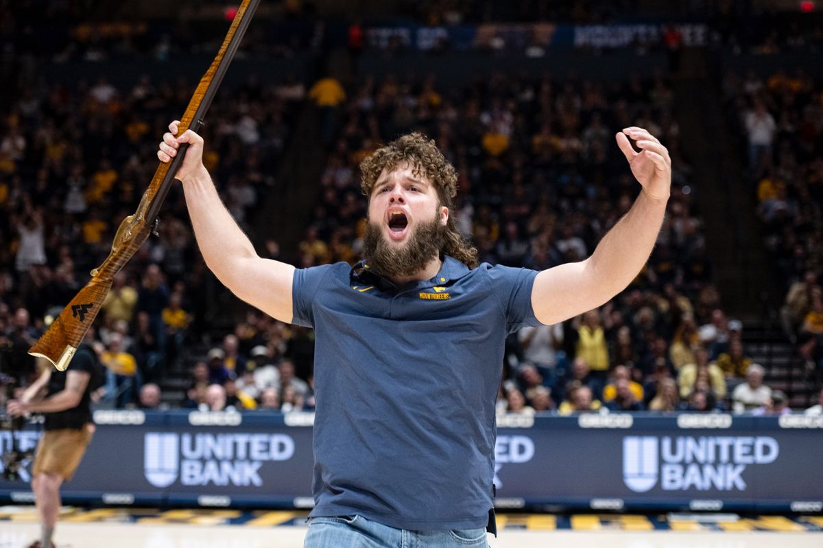 Mikel Hager celebrates on the floor of the Coliseum after being named the Mountaineer mascot. He is wearing a blue shirt and is holding the rifle in his right hand, cheering for the crowd.
