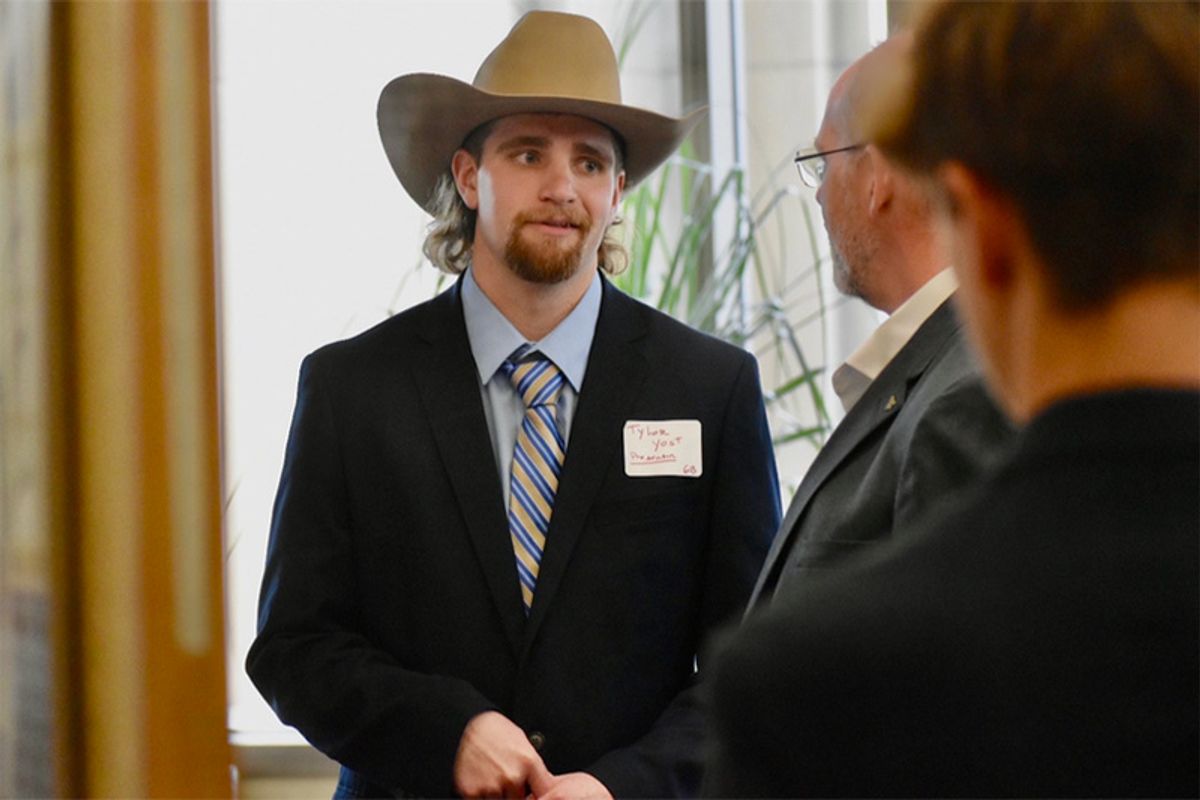 Photograph of WVU student Tylor Yost wearing a tan cowboy hat and a black coat over a light blue button up shirt and striped tie. He is standing with two other men whose faces you cannot see. 