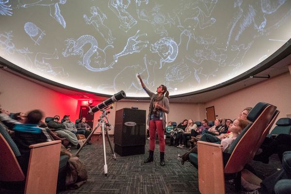Photograph of an expert pointing up towards the illuminated ceiling of the WVU planetarium. There are people seated in chairs around her and a telescope standing near her. 