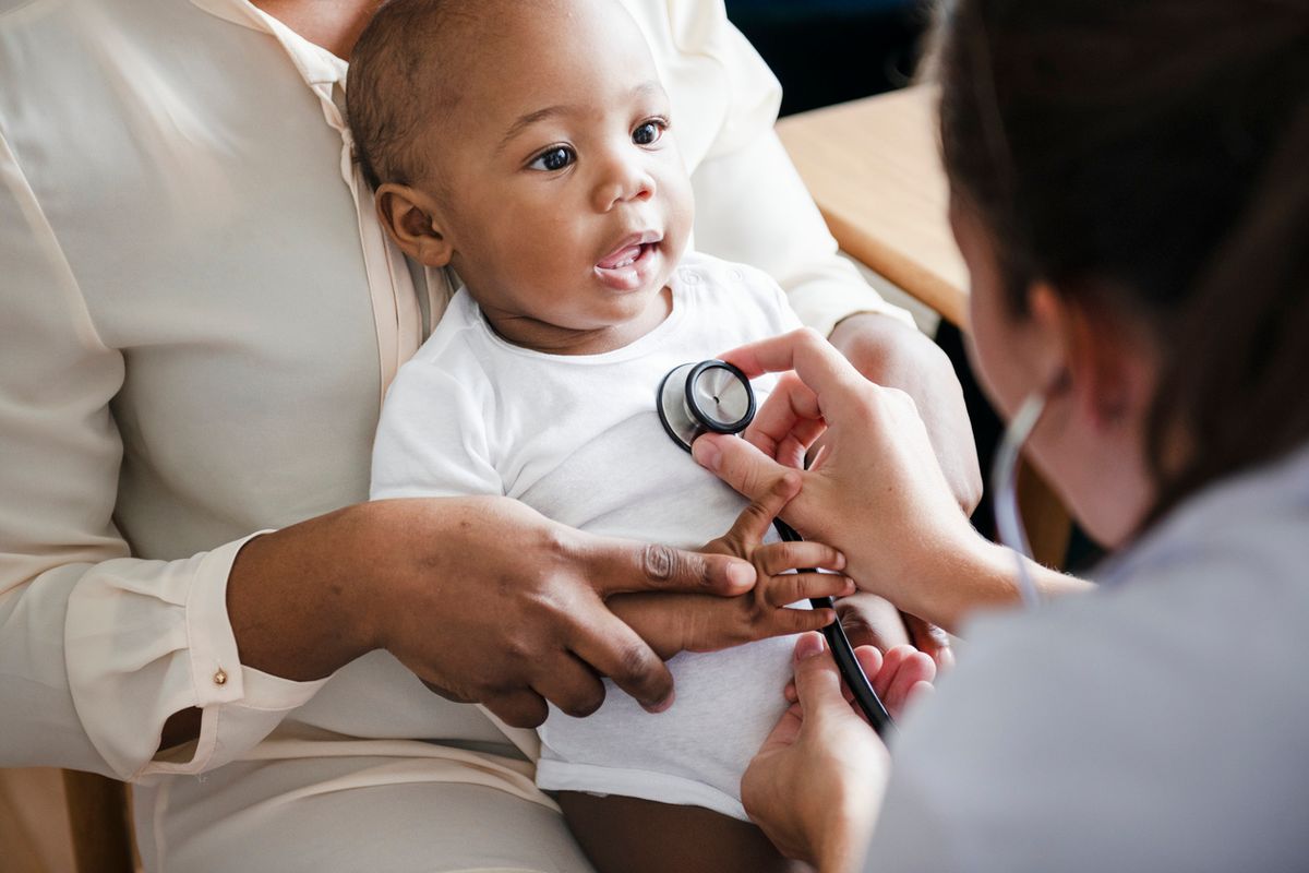 A woman in a white blouse (face not shown) holds a baby with dark brown eyes in her lap while a doctor uses a stethoscope on the baby. 