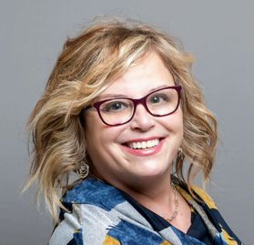 WVU Professor Kim Floyd. She is pictured against a gray background. She has wavy, shoulder-length blond hair and is wearing a blue, gray and gold patterned blouse. She is smiling and is wearing red-framed glasses. 