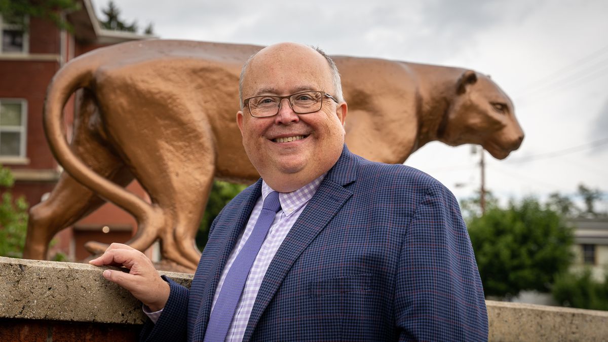 Photo of WVU Potomac State College incoming president Chris Gilmer. He is standing in front of a bronze statue of a mountain lion, the school's mascot. He is wearing a navy blue suit with a purple tie and glasses. 
