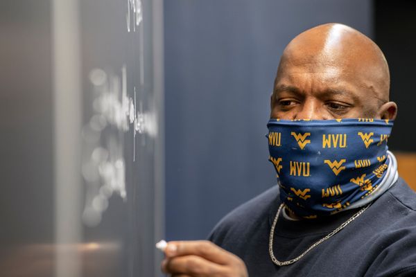 A man in a WVU gold and blue face covering writes on a black board with chalk