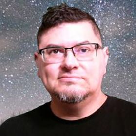 Headshot of WVU professor Jason Ybarra. He is pictured in front of a space background and is wearing a black shirt and square framed glasses. He has brown hair and a short gray goat tee. 