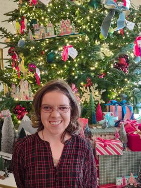 Photograph of WVU employee Mary Veselicky standing in front of a large, decorated Christmas tree. Mary is wearing a red plaid blouse and has shoulder length light brown hair. 