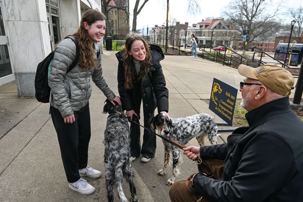 Three people are outside the Mountainlair. On the right, a person with a tan hat and black jacket holds two English setters on a leash. Two people pet the dogs. They are wearing gray and black puffer jackets and have on backpacks.