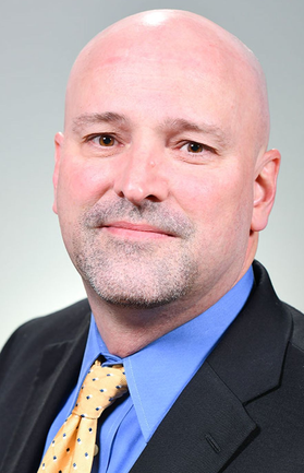 Headshot of WVU administrator Brice Knotts. He is pictured in front of a light gray background wearing a dark suit jacket over a blue shirt and gold patterned tie. His head is shaved and he has light facial hair. 