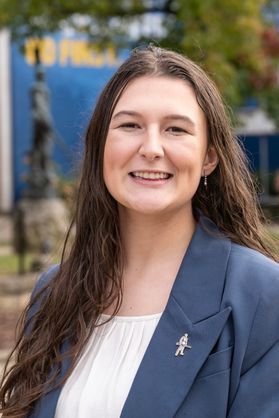 Headshot of WVU student Olivia Dowler. She is pictured outside wearing a navy blue suit jacket over a white pleated blouse. She has long brown hair. 