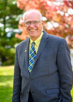 Headshot of WVU honoree Bob Wanker. He is pictured outside and is wearing a yellow dress shirt with a gold and blue striped tie under a blue plaid sport coat. He has receding white hair and wears glasses. 