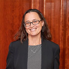 Headshot of WVU researcher Maura McLaughlin. She is standing in front of a wall of cherry wood paneling and is wearing a black jacket over a gray shirt. She has long brown hair and wears black, square-framed glasses. 