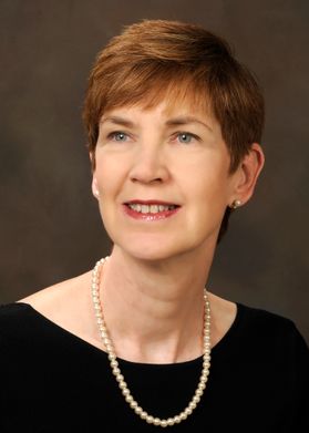 Headshot of WVU honoree Nora MacDonald. She is pictured against a dark colored background wearing a black blouse and a string of pearls. She has short red hair. 