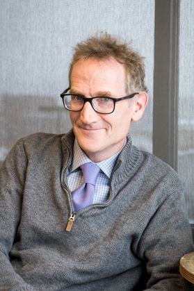 Man in glasses wearing a grey sweater and purple tie sits and smiles for picture 