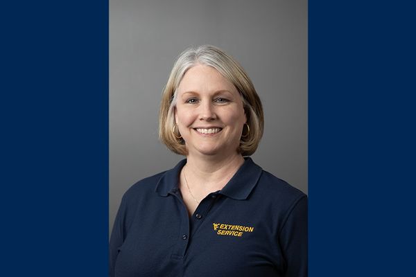 Image with a headshot of WVU Extension agent Gwen Crum wearing a branded, navy blue golf shirt posed against a gray background. She has shoulder length light colored hair. The headshot is centered on a blue background. 