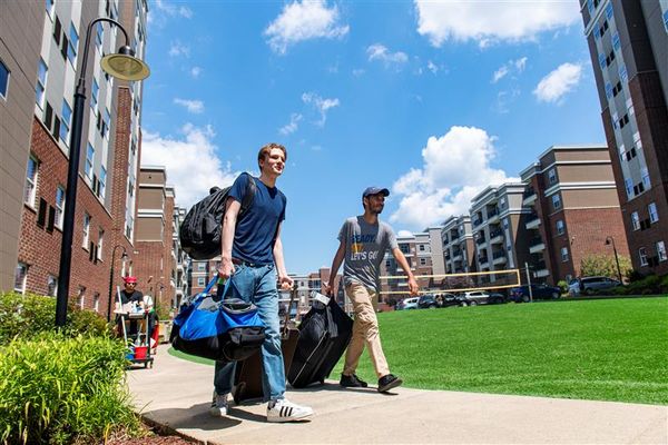 WVU students move into residence halls during the fall semester. A male student wearing a blue T-shirt and blue jeans walks near residence halls holding multiple bags. A male WVU volunteer wearing a WVU Tshirt and khaki pants walks with him rolling a bag.