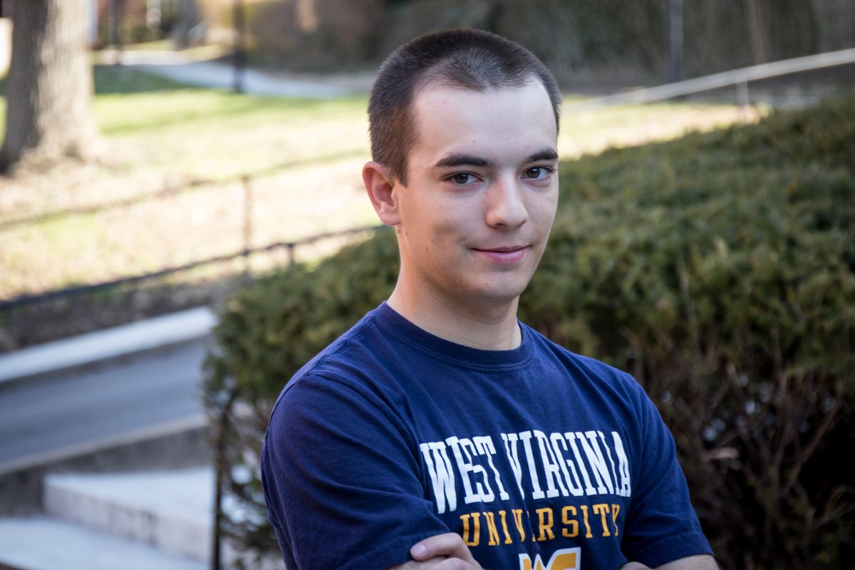 photo of young man with short dark hair wearing West Virginia University shirt, outside in front of shrubbery