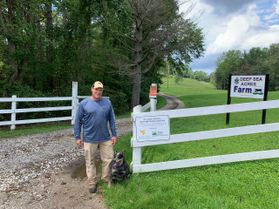 Kevin Jones, owner of Deep Sea Acres Farm in Kanawha County, is working with WVU researchers on his 250-acre farm. He is pictured standing near a white fence that marks the entrance of his farm. There is a sign that is barely legible to his right. 