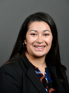 Headshot of scholarship recipient Paola Perez-Vega. She is pictured in front of a gray background and is wearing a black jacket over a colorful patterned shirt. She has long black hair.